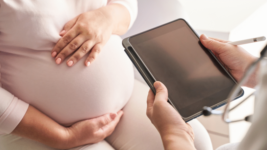 A pregnant woman places her hand on her belly while consulting with a doctor holding a tablet during a prenatal visit.