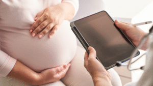 A pregnant woman places her hand on her belly while consulting with a doctor holding a tablet during a prenatal visit.