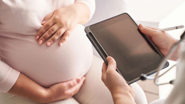 A pregnant woman places her hand on her belly while consulting with a doctor holding a tablet during a prenatal visit.